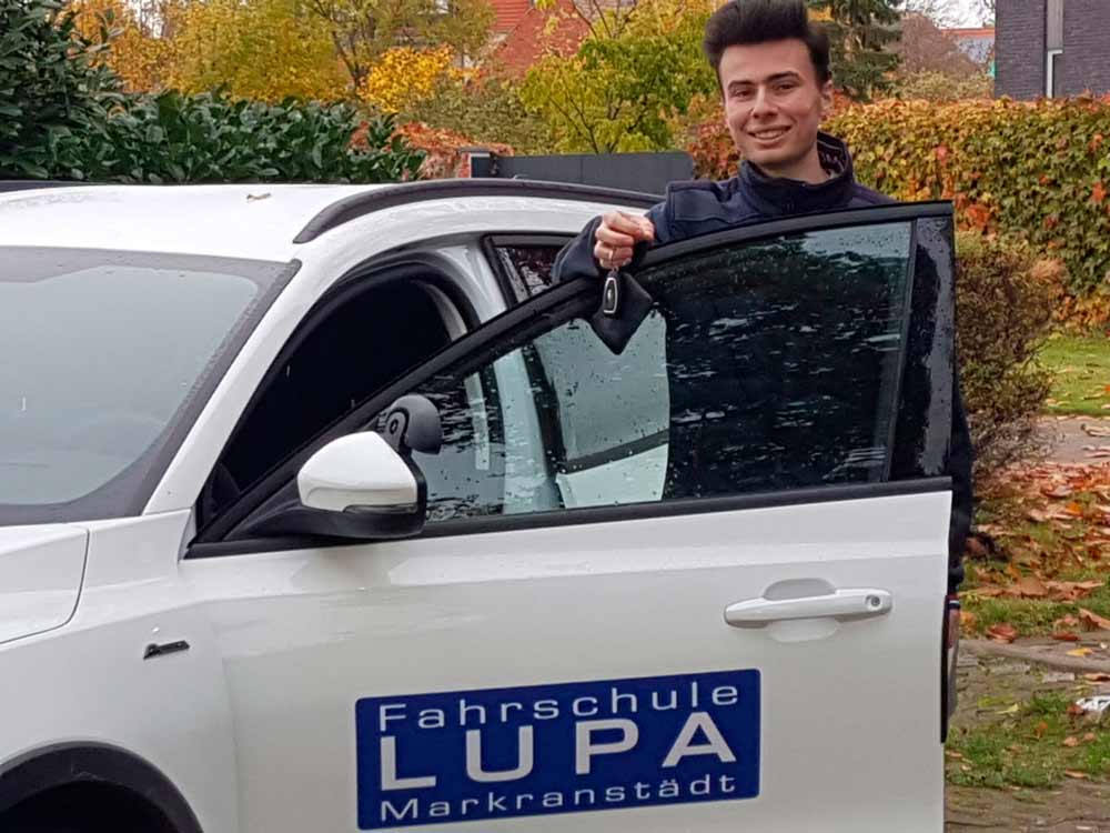 Junger Mann steht am offenen Auto mit einem Autoschlüssel in der Hand und lächelt in die Kamera, weil er die Fahrprüfung bestanden hat.